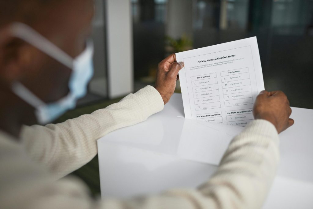 Afroamerikanischer Mann mit Gesichtsmaske, der mit offiziellem Stimmzettel seine Stimme abgibt