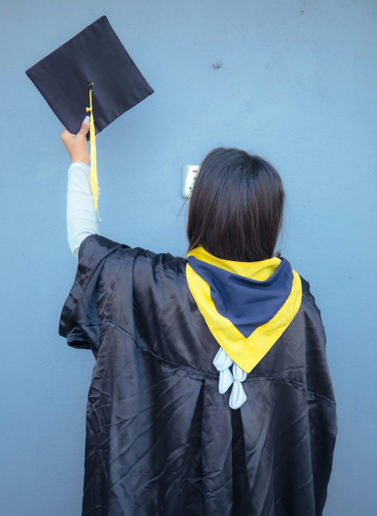 eine Frau in einem Abschlusskleid hält einen schwarzen Abschlusshut hoch